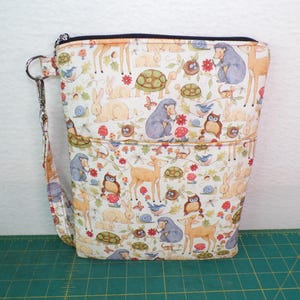 May include: A rectangular zippered pouch with a wrist strap featuring a repeating pattern of woodland animals including deer, rabbits, hedgehogs, turtles, and owls, interspersed with small red mushrooms, blue birds, and snails on a cream background.