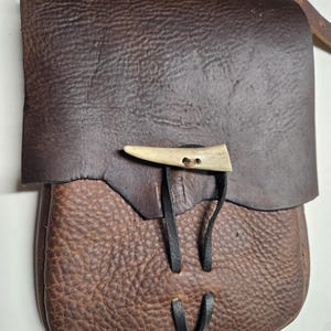 May include: A dark brown leather pouch featuring a textured surface and a flap closure. The bag is secured with a light-coloured, curved bone toggle button and a thin black leather cord that loops through the flap and the main body of the bag.