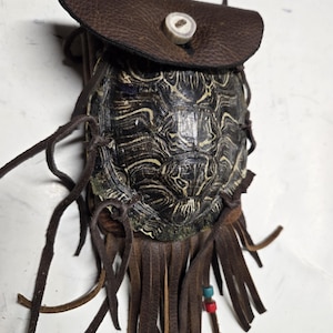 May include: A handcrafted leather pouch with a turtle shell base. The pouch features a dark brown leather flap secured with a button, and a fringe of leather strips. Small beads in red and blue add a pop of color.