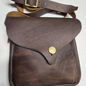 May include: A dark brown leather crossbody bag with a textured surface, a fold-over flap, and a single brass-colored snap button closure. The bag features an adjustable leather shoulder strap with a rectangular brass-colored metal buckle.