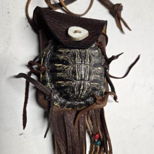 May include: A brown leather pouch featuring a dark turtle shell on the front flap, secured with a white bone button. The pouch includes long leather fringe tassels at the bottom, accented with small red and blue beads, and a leather neck strap.