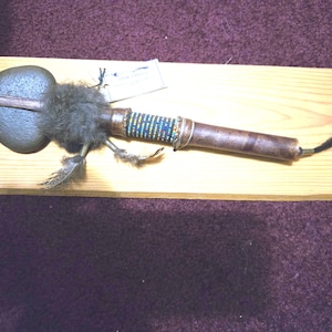 May include: A Native American ceremonial object with a rounded stone head and a wooden handle. The handle is decorated with beads, feathers, and fur. A black tassel hangs from the end. The object rests on a wooden plank.