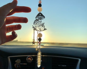 May include: A silver car charm featuring a mushroom design with crystals and beads hanging from it. The charm is hanging from a rearview mirror in a car.