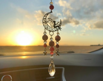Puede incluir: Un amuleto de coche de metal negro en forma de media luna con una silueta de hada sentada sobre él. El amuleto está adornado con cuentas rojas y transparentes y un colgante de cristal en forma de lágrima. El amuleto está colgado de un espejo retrovisor en un coche.