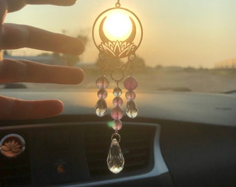 Puede incluir: Un amuleto para el coche con un diseño de luna creciente y flor de loto plateada con cuentas de cristal morado y transparente que cuelgan de él. El amuleto está colgado del espejo retrovisor de un coche.