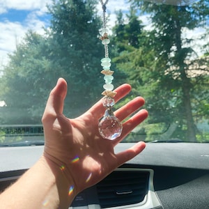 May include: A crystal car charm with a silver metal top and a beaded chain with light blue and white beads. The charm is hanging from a rearview mirror in a car.