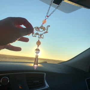 May include: A gold elephant charm with pink and white beads hangs from a rearview mirror. A crystal prism dangles from the bottom of the charm.