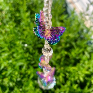 Op de afbeelding: Een kristallen zonenvanger met iriserende vlinders die aan een kralenketting hangen. De vlinders zijn paars, blauw en groen.