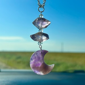 May include: A silver chain with three amethyst crystals and a crescent moon-shaped amethyst charm. The charm is purple and has a light purple hue.