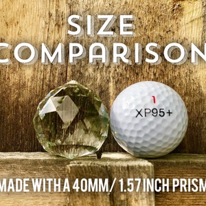 Op de afbeelding: Groottevergelijking van een 40 mm / 1,57 inch kristallen prisma naast een golfbal. De golfbal heeft de tekst "XP95+" erop gedrukt.