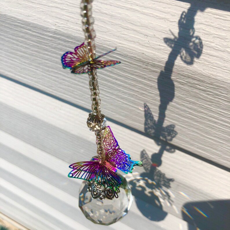 Op de afbeelding: Een kristallen zonenvanger met iriserende vlinders die aan een kralenketting hangen. De zonenvanger is helder en heeft een facetgeslepen ontwerp. De vlinders zijn van metaal en hebben een regenboogkleurige afwerking.