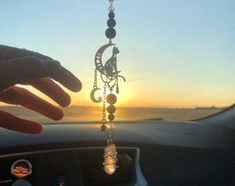Puede incluir: Un amuleto de coche plateado con una luna creciente, un gato sentado en la luna y un cristal colgando de la parte inferior. El amuleto está colgado de un espejo retrovisor en un coche. El sol se está poniendo en el fondo.