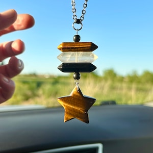 May include: A gold star-shaped charm with tiger's eye stone hangs from a silver chain. The charm is attached to three stacked rectangular stones: brown tiger's eye, clear quartz, and black onyx.