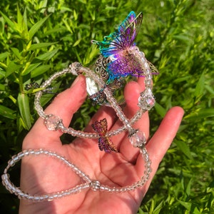 Op de afbeelding: Een ketting van heldere kristallen kralen met iriserende vlinderbedels. De ketting is geregen met kleine, heldere kralen en heeft verschillende vlinderbedels met iriserende vleugels.