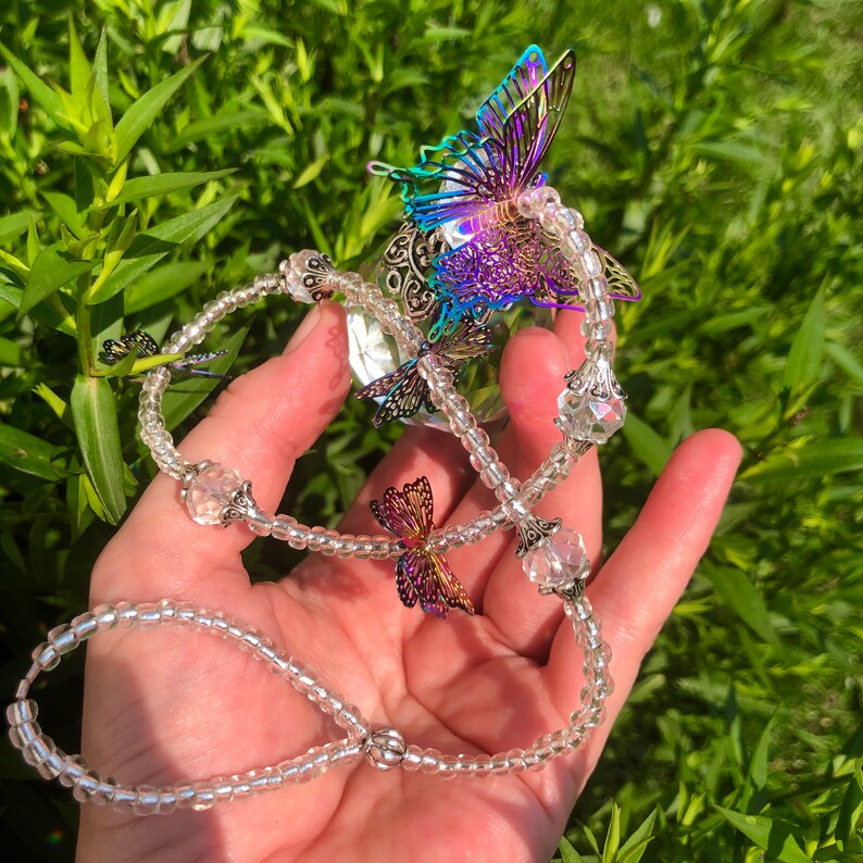 Op de afbeelding: Een ketting van heldere kristallen kralen met iriserende vlinderbedels. De ketting is geregen met kleine, heldere kralen en heeft verschillende vlinderbedels met iriserende vleugels.