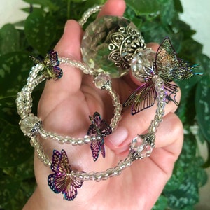 Op de afbeelding: Een kristallen bolbedel met een zilveren metalen ontwerp en iriserende vlinderaccenten hangt aan een heldere kralenketting.