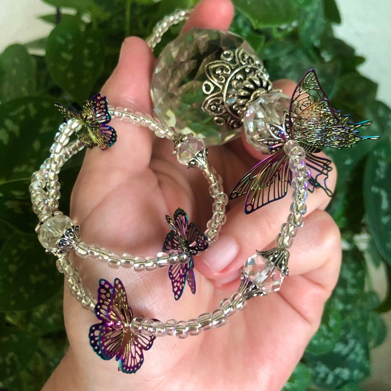 Op de afbeelding: Een kristallen bolbedel met een zilveren metalen ontwerp en iriserende vlinderaccenten hangt aan een heldere kralenketting.