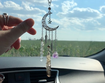 Puede incluir: Un amuleto de coche de plata en forma de media luna con una estrella y cristales colgantes. Los cristales son transparentes, marrones y morados.