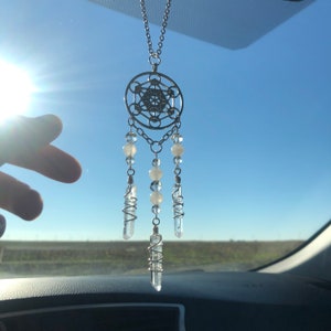 May include: A silver car charm with a geometric design and clear crystal beads hanging from it. The charm is hanging from a rearview mirror in a car.