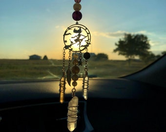May include: A silver car charm with a witch silhouette and crystals hanging from it. The charm is hanging from the rearview mirror of a car. The sun is setting in the background.