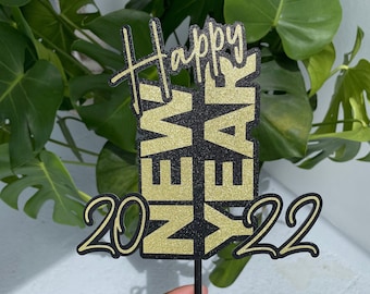 Op de afbeelding: Een zwarte en gouden glittertaarttopper met de woorden "Happy New Year" en de cijfers "2022".