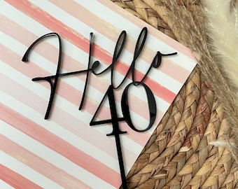 Op de afbeelding: Zwarte acryl taarttopper met de woorden "Hello 40" in een handschrift. De topper is op een roze en witte gestreepte achtergrond.