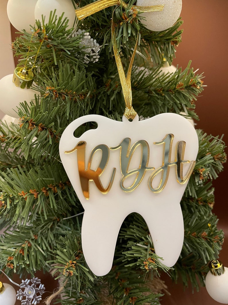 Op de afbeelding: Witte, tandenvormige kerstversiering met een gouden glitterafwerking en de naam "Ryff" in gouden letters.
