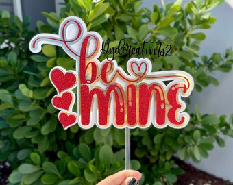Op de afbeelding: Een witte taarttopper met de woorden "be mine" in rode glitterletters met een gouden rand. Aan de linkerkant van de topper zitten drie rode glitterhartjes.