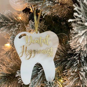 Op de afbeelding: Witte, tanden-vormige kerstversiering met gouden glitter-lettering die "Dental Hygienist" zegt.