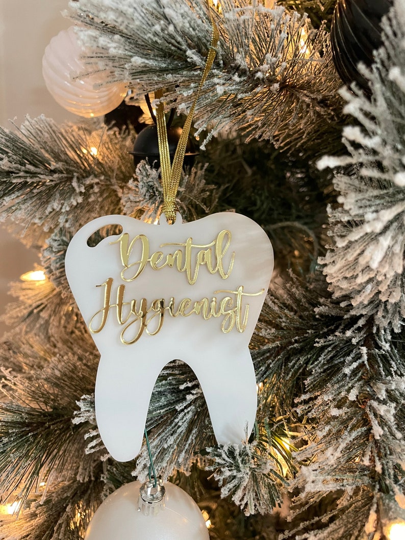 Op de afbeelding: Witte, tanden-vormige kerstversiering met gouden glitter-lettering die "Dental Hygienist" zegt.
