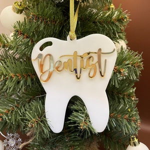 Op de afbeelding: Witte, tandenvormige kerstversiering met het woord "Dentist" in gouden glitterletters.
