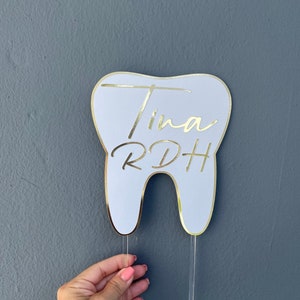 May include: White tooth-shaped cake topper with gold trim and the text "Tina RDH" in gold lettering.