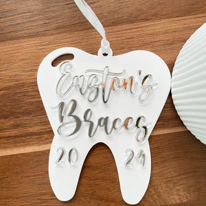 Op de afbeelding: Witte, tandenvormige hanger met de tekst "Easton's Braces 20 24" in zilveren letters.