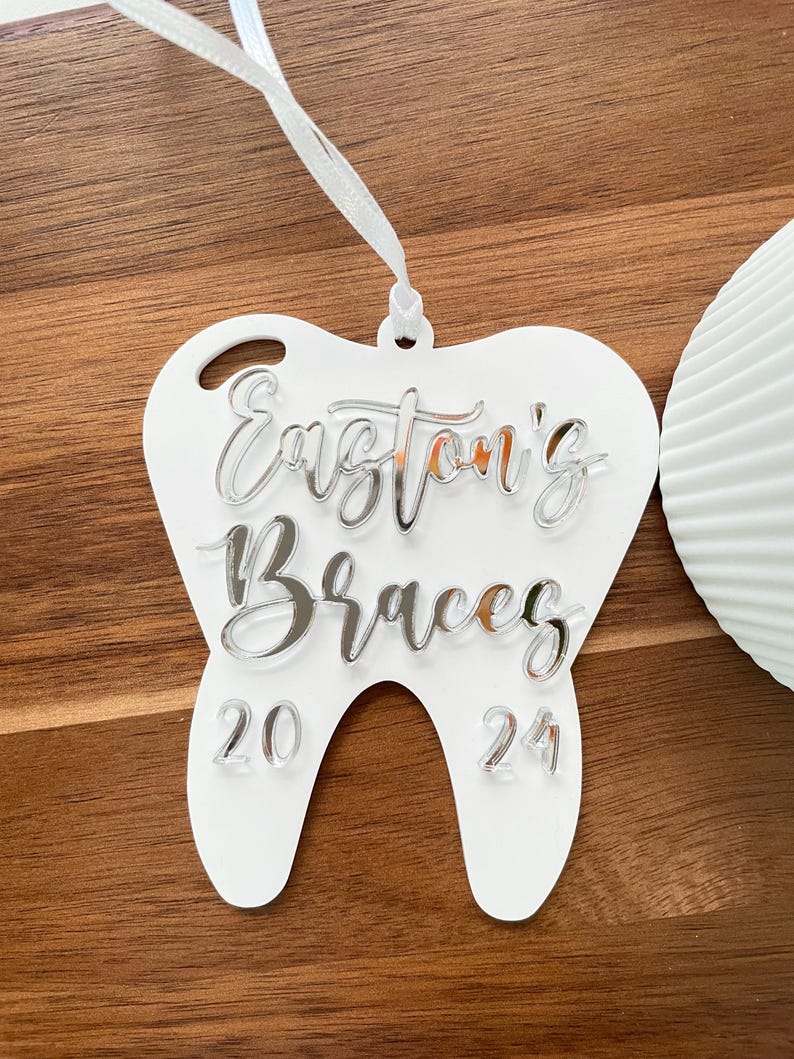 Op de afbeelding: Witte, tandenvormige hanger met de tekst "Easton's Braces 20 24" in zilveren letters.