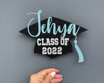 Op de afbeelding: Zwarte afstudeerhoed-taarttopper met zilveren glittertekst die "Jehya Class of 2022" luidt. De hoed heeft een blauwe glitterkwast.