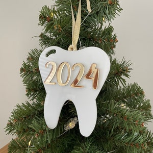 Op de afbeelding: Witte kerstversiering in de vorm van een tand met gouden 2024-lettering hangend aan een groene kunstkerstboom.