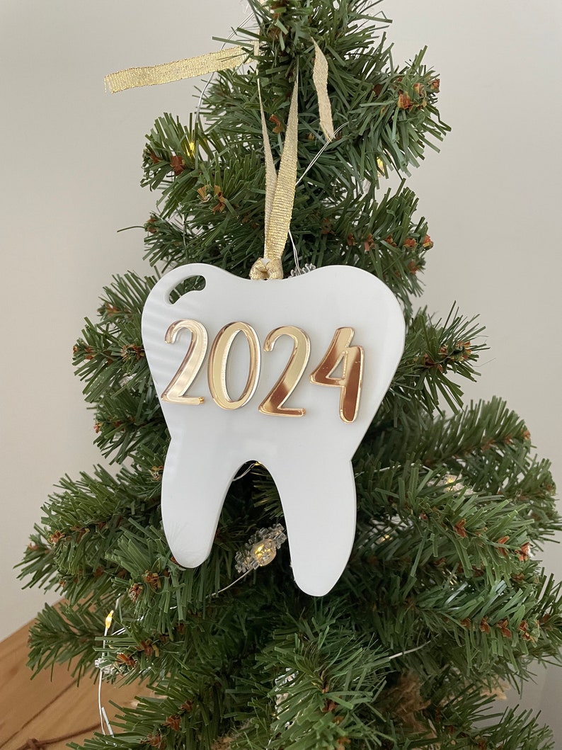 Op de afbeelding: Witte kerstversiering in de vorm van een tand met gouden 2024-lettering hangend aan een groene kunstkerstboom.