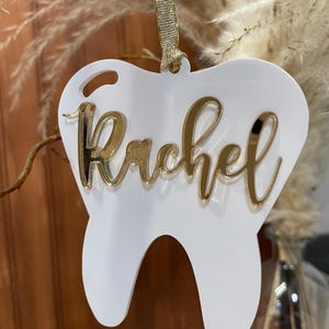Op de afbeelding: Witte, tandenvormige hanger met een gouden glitterafwerking en de naam "Rachel" in gouden glitterletters.