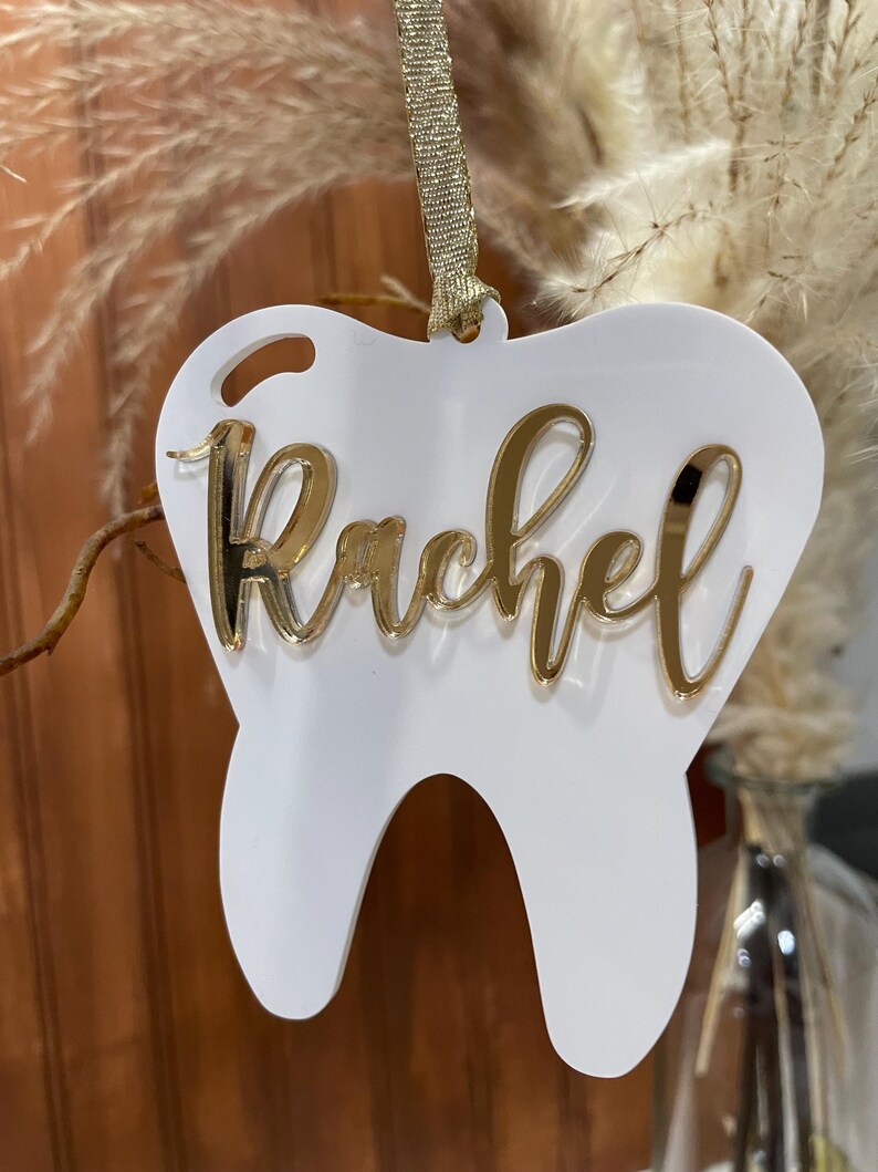 Op de afbeelding: Witte, tandenvormige hanger met een gouden glitterafwerking en de naam "Rachel" in gouden glitterletters.