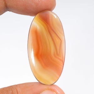 May include: An oval-shaped cabochon gemstone featuring a smooth, polished surface with translucent layers of orange, amber, and cream-colored banding. The stone displays a natural, swirling pattern of warm earth tones throughout its polished face.
