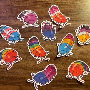 Puede incluir: Un conjunto de diez pegatinas de cochinillas coloridas con diseños de banderas del arcoíris, transgénero y bisexual. Cada pegatina presenta una cochinilla de dibujos animados con un corazón.