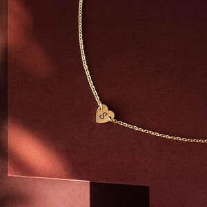May include: A gold chain necklace with a small heart pendant. The heart has the letter "S" engraved on it. The necklace is photographed on a burgundy background.