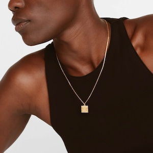 Puede incluir: Una persona lleva un top negro de tirantes y un collar de cadena dorada con un pequeño colgante cuadrado.