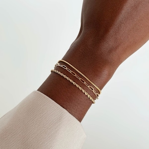 以下が含まれることがあります： さまざまなスタイルの3つのゴールドブレスレットが手首に重ねられています。ブレスレットには、ロープチェーン、ペーパークリップチェーン、スネークチェーンが含まれます。その人はベージュの長袖シャツを着ています。