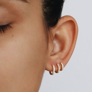 Può includere: Primo piano di un orecchio con tre orecchini a cerchio d'oro con piccole pietre di zircone cubico trasparenti.