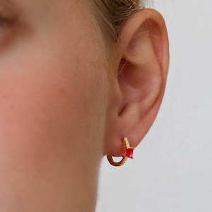 Op de afbeelding: Gouden creolen met een klein rood vierkant steentje. De oorbellen worden in het oor van een persoon gedragen.
