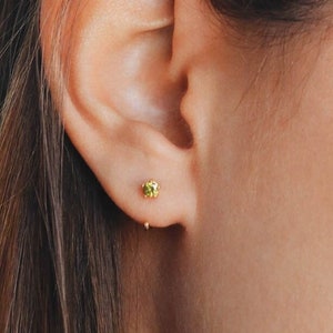 Puede incluir: Primer plano de una oreja con un pequeño pendiente de piedra preciosa verde. El pendiente es un simple botón con un engaste de oro.