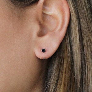 Könnte beinhalten: Nahaufnahme eines Ohrs mit einem kleinen schwarzen Edelstein-Ohrstecker. Der Ohrring ist in einer einfachen goldenen Fassung gefasst.