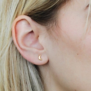 Op de afbeelding: Een close-up van een oor met een kleine, gouden halve maan oorbel. De oorbel heeft een minimalistisch ontwerp. De persoon heeft blond haar en een lichte huid. De achtergrond is een neutrale kleur.