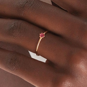 Op de afbeelding: Een delicate gouden ring met een kleine, ronde, rode edelsteen. De ring heeft een dunne band en een eenvoudig, elegant ontwerp. De edelsteen is gezet in een klassieke prong setting, wat een vleugje kleur en sprankeling toevoegt.